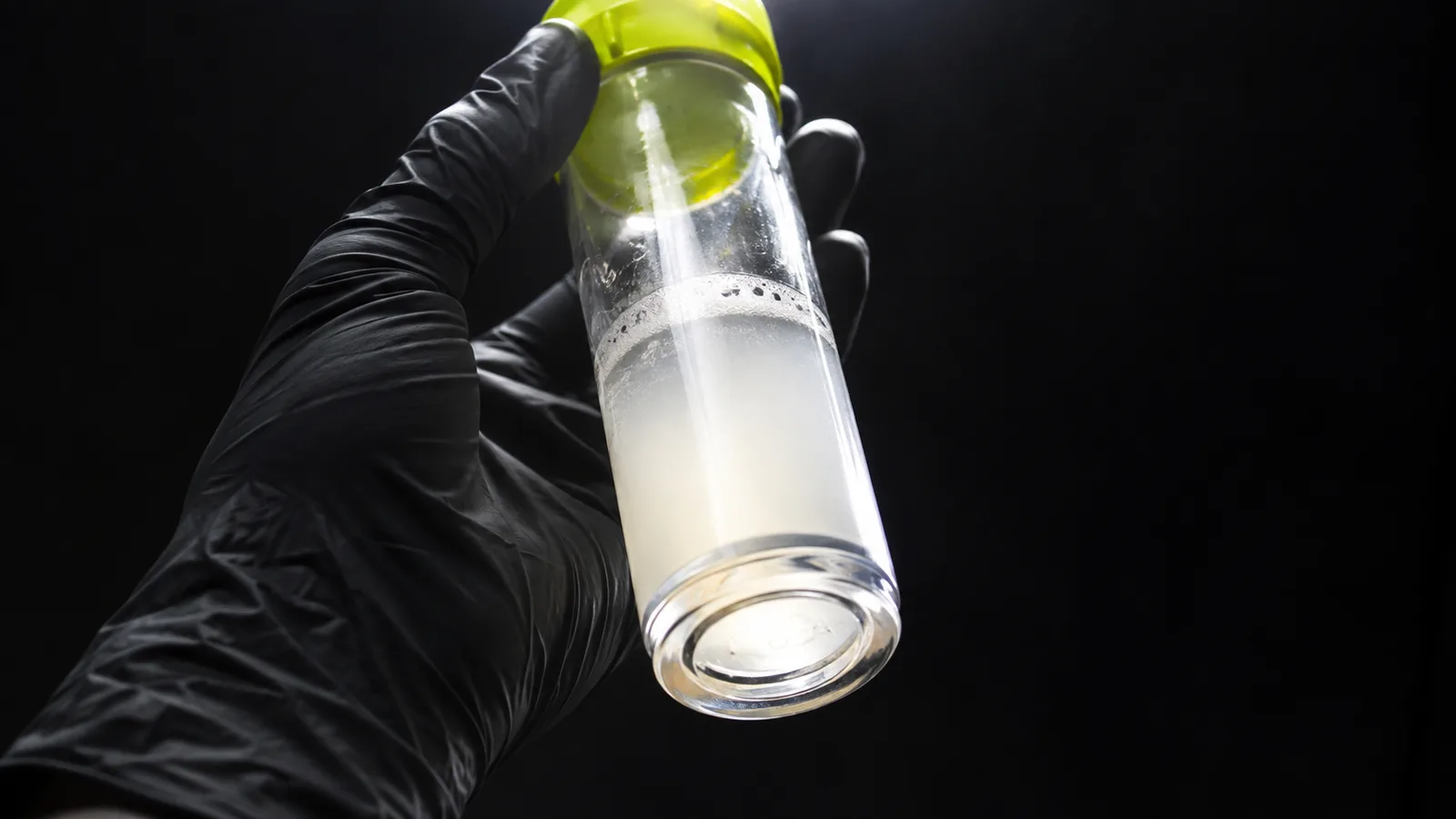 A black-gloved hand holds up a clear canister of cloudy collected fluid against a black background.